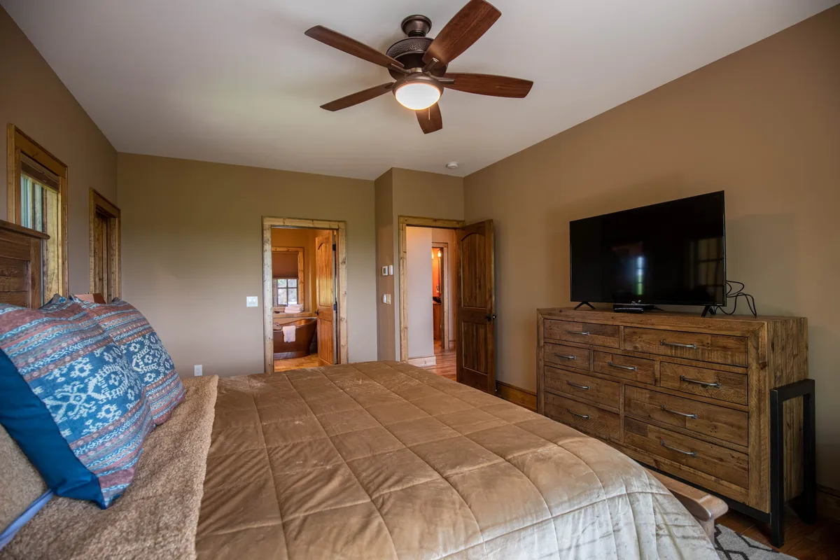 A spacious bedroom with a large bed dressed in plush linens, a ceiling fan overhead, a flat screen TV mounted on the wall, a dresser, and soft lighting that creates an inviting and restful atmosphere. The room feels airy and comfortable, designed for relaxation and quiet evenings in.