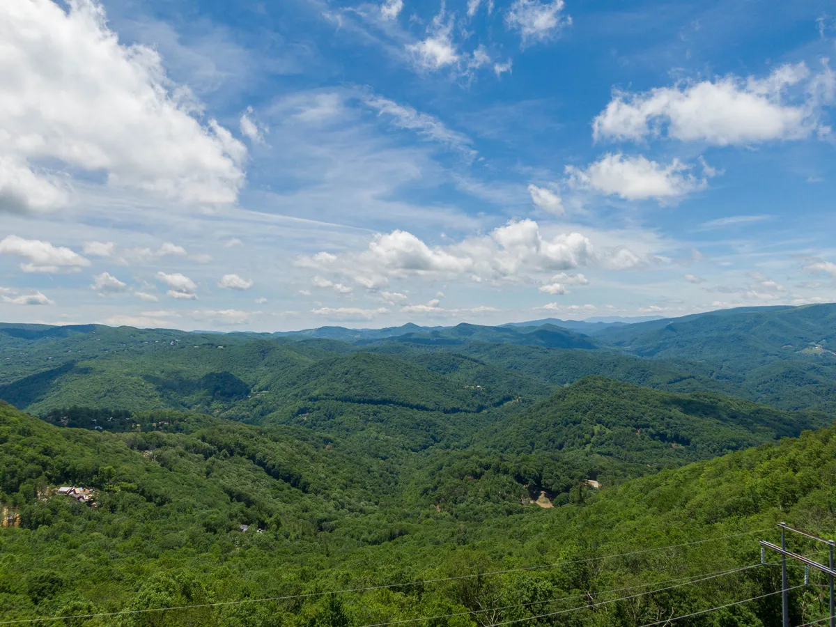 A sweeping view of mountain ranges and lush green forests under a blue sky with scattered clouds. The scene suggests expansive trails and natural beauty, making it ideal for outdoor activities such as hiking, nature walks, and bird watching.