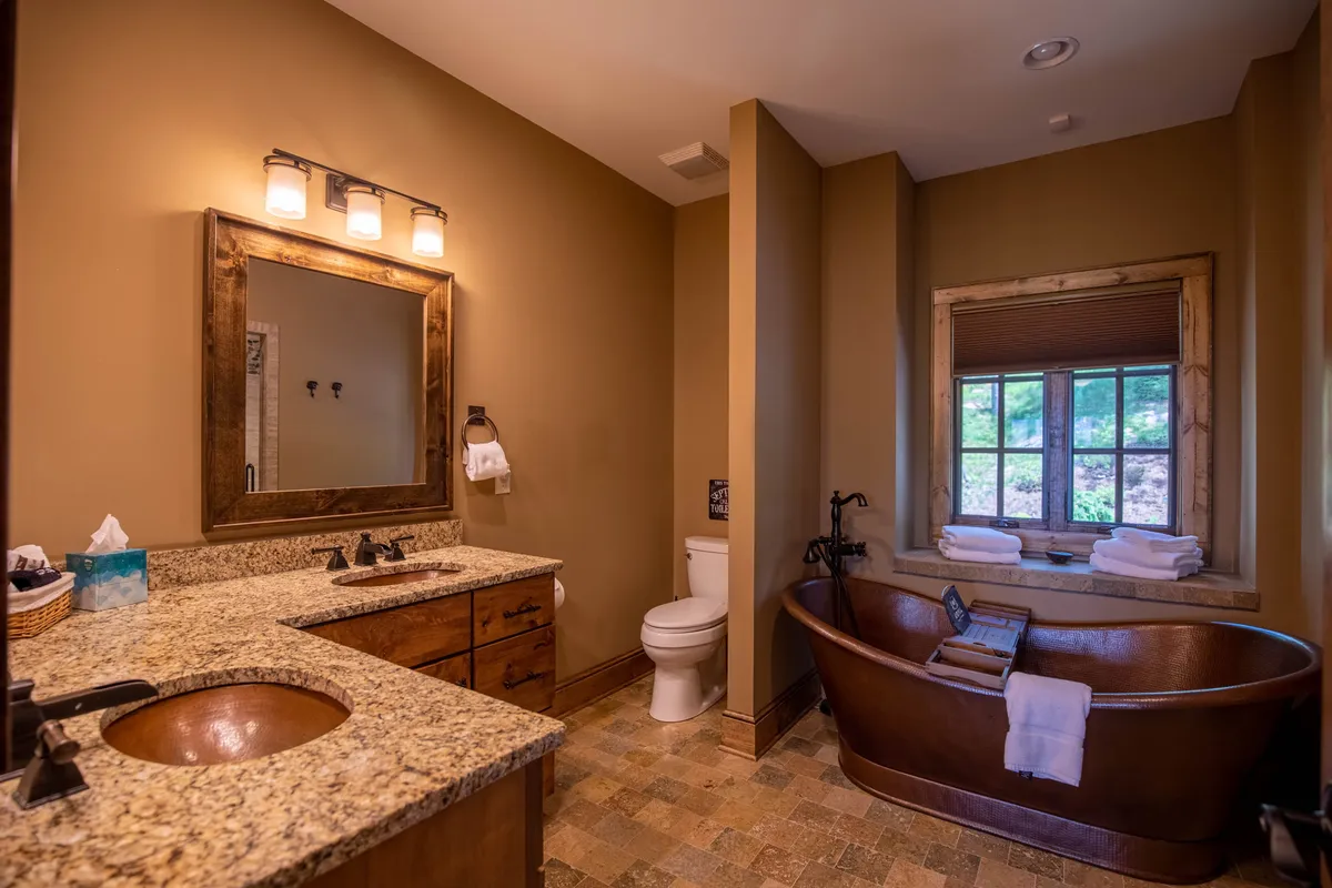 A serene bathroom space featuring a freestanding tub with hammered copper accents, a granite countertop vanity with two sinks, a large mirror, and a window that lets in natural light. The overall ambiance is spa-like and indulgent, offering a peaceful retreat for self-care and relaxation.