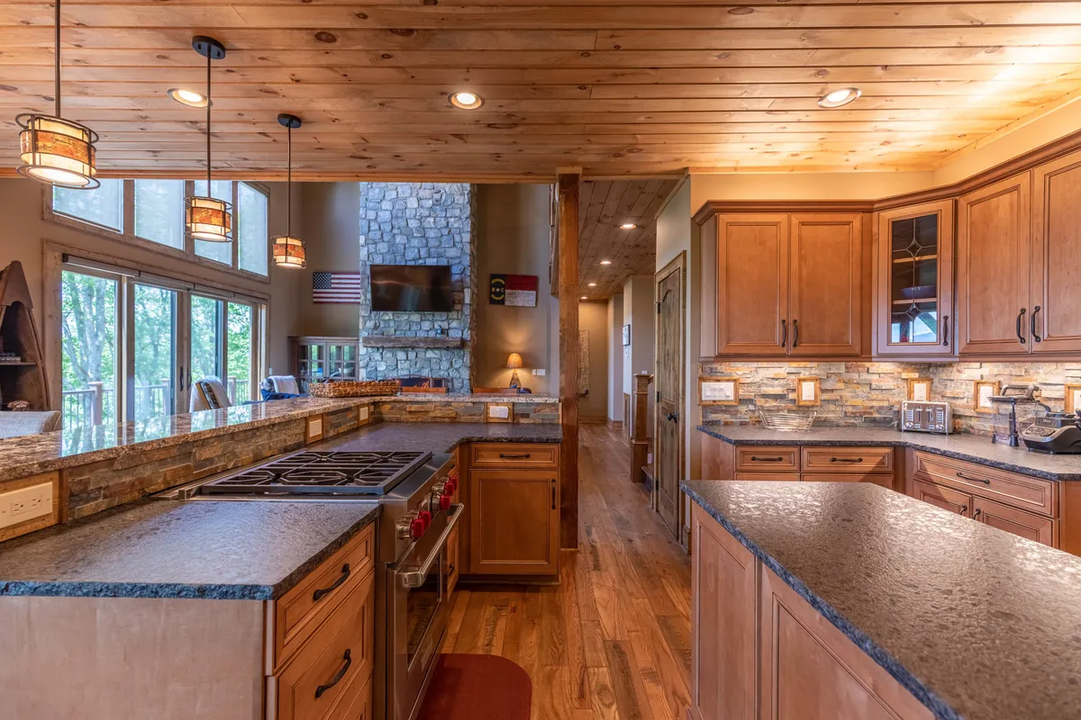 An open-plan layout uniting the kitchen, dining, and living areas. The kitchen island with stovetop sits adjacent to a stone fireplace, with large windows letting in natural light. The arrangement encourages gatherings, meal prep, and easy socializing across spaces.