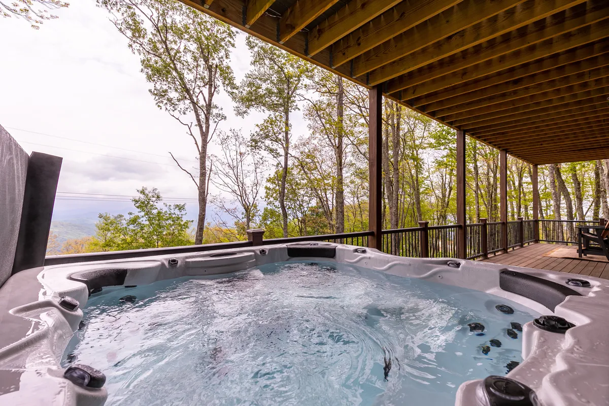 An inviting exterior deck featuring a bubbling hot tub surrounded by lush trees, with serene mountain views in the background. The hot tub is the focal point, offering a space to relax outdoors after a day of adventure.