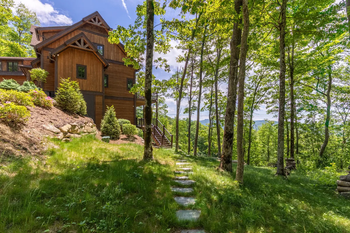 The exterior of a luxury wooden house nestled among lush greenery and trees, with a stone pathway leading to the entrance. The tranquil garden and forested surroundings evoke privacy and the promise of seasonal outdoor activities.