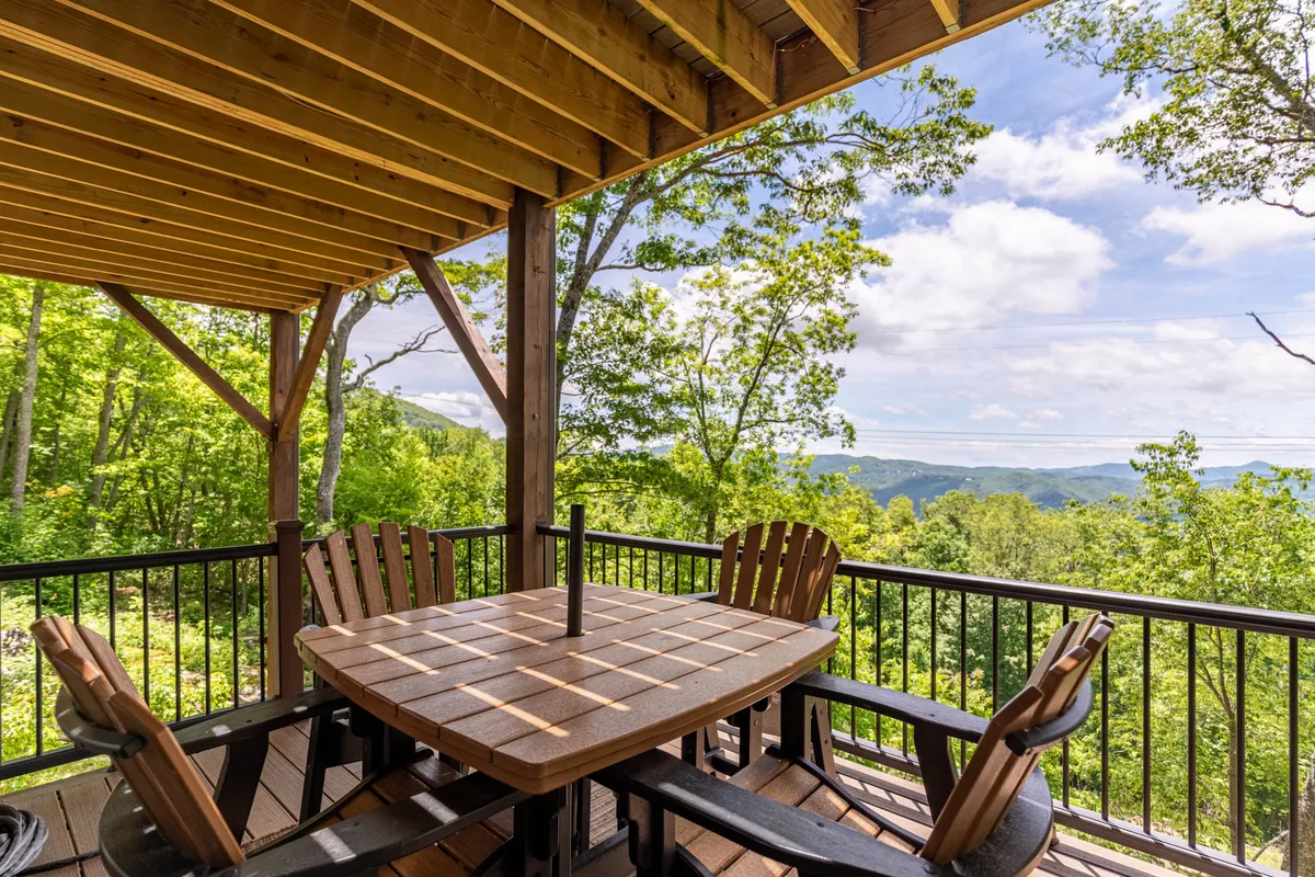 An outdoor dining area with a wooden table and Adirondack chairs set against a backdrop of distant mountains and lush greenery. The tranquil setting invites socializing, dining, and soaking in the mountain views together.