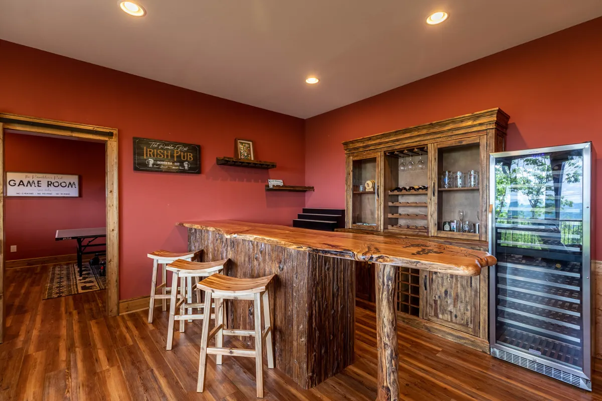 A custom wooden bar with bar stools, a wine cooler, and shelves lined with glassware. The cozy setting is designed for socializing, tasting beverages, and playing games, inviting guests to linger and enjoy each other's company.