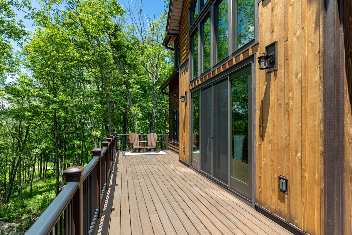 A spacious wooden deck with comfortable Adirondack chairs overlooks lush greenery. Large windows line the house, blending indoor and outdoor spaces. The serene natural setting is perfect for relaxation and taking in the views.