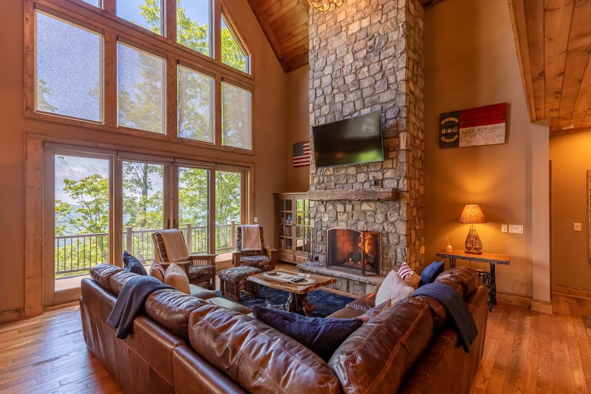 A spacious great room featuring a large stone fireplace as the focal point, complemented by a leather sectional sofa and a wooden coffee table. Floor-to-ceiling windows fill the space with natural light and offer sweeping views of the outdoors, while the vaulted ceiling adds grandeur to the room.